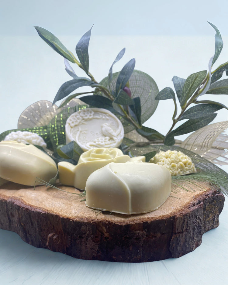 Close-up of Nardus Bliss artisanal soap—cream‑toned bar shaped as a rose, positioned on a sliced olive‑wood slab, with dried lavender buds, olive sprig foliage, and rustic ribbon in the background for a Greek‑Mediterranean aesthetic. DEOS CO.