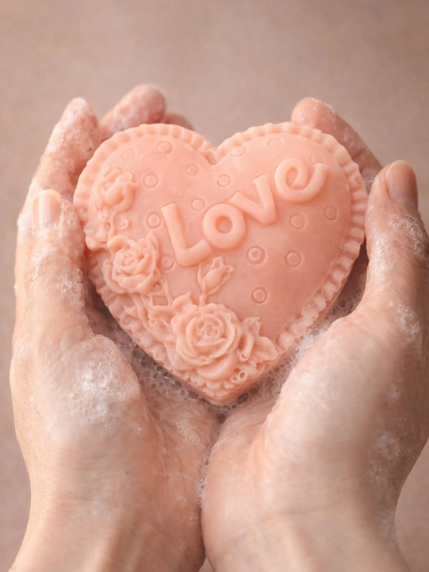 Heart-shaped soap with 'LOVE' and floral designs held between hands against a beige background by DEOS CO. Valentine's Day rose geranium and vanilla soap