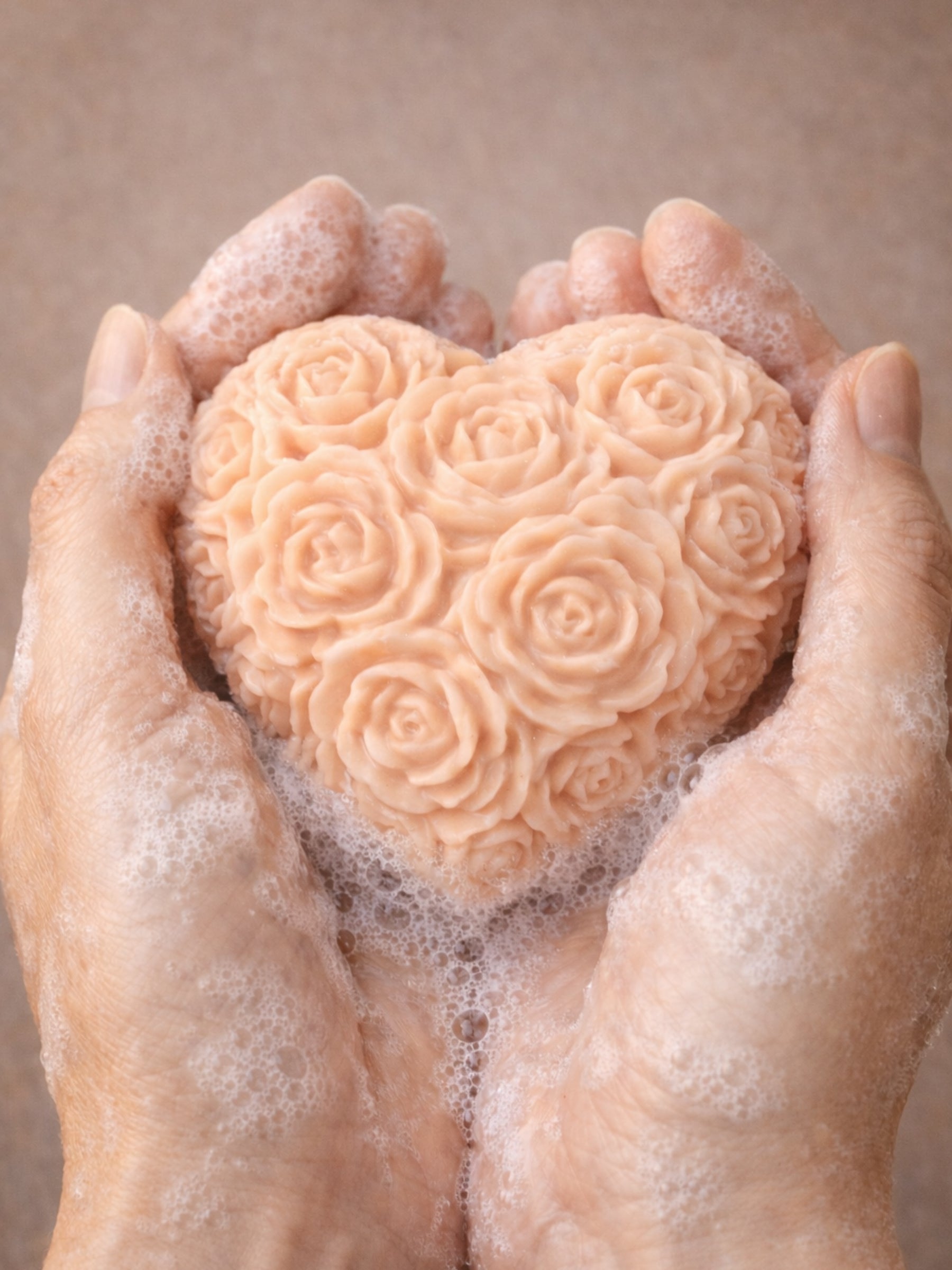 Hands holding the Fleur Heart Grand Bloom Valentine’s Day soap with soft lather, highlighting its fuller heart shape. Made of greek organic olive oil, rose geranium oil, mango oil, vanilla essential oil, coconut oil