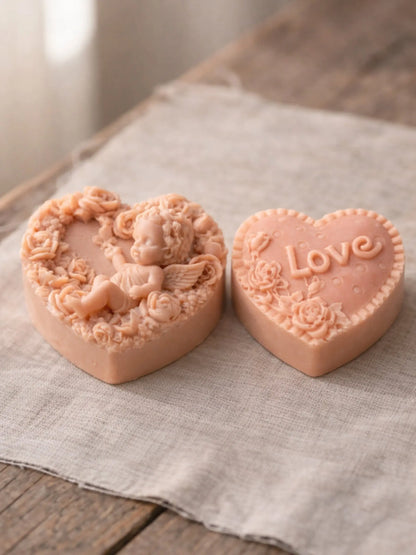 Two heart-shaped peach pink soap bars with angel and 'Love' design on a textured fabric background DEOS CO soap orgnaic olive oil soap with rose geranium and vanilla. Eros Angel and LOVE, DEOS soaps