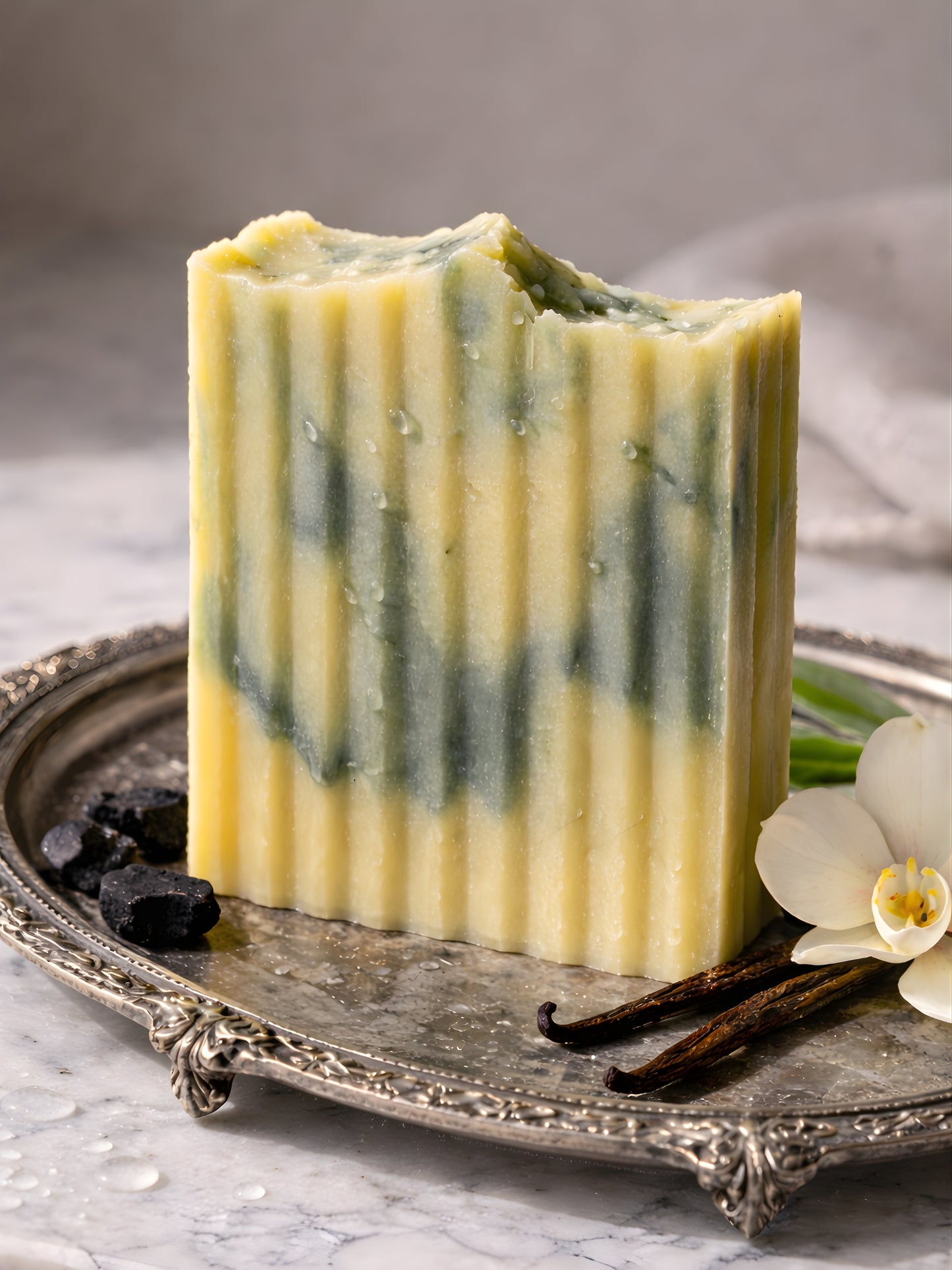 Bar of soap with green and yellow stripes on a decorative tray with vanilla beans and flowers.