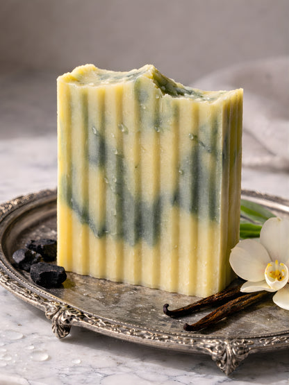 Bar of soap with green and yellow stripes on a decorative tray with vanilla beans and flowers.