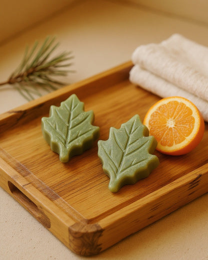 Two leaf-shaped green soap bars on a wooden tray with an orange and a towel in the background.