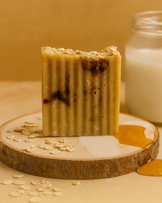 Bar of natural organic soap with oatmeal pieces, honey and spilled oatmilk on a wooden board with a jar in the background by DEOS Co. brand