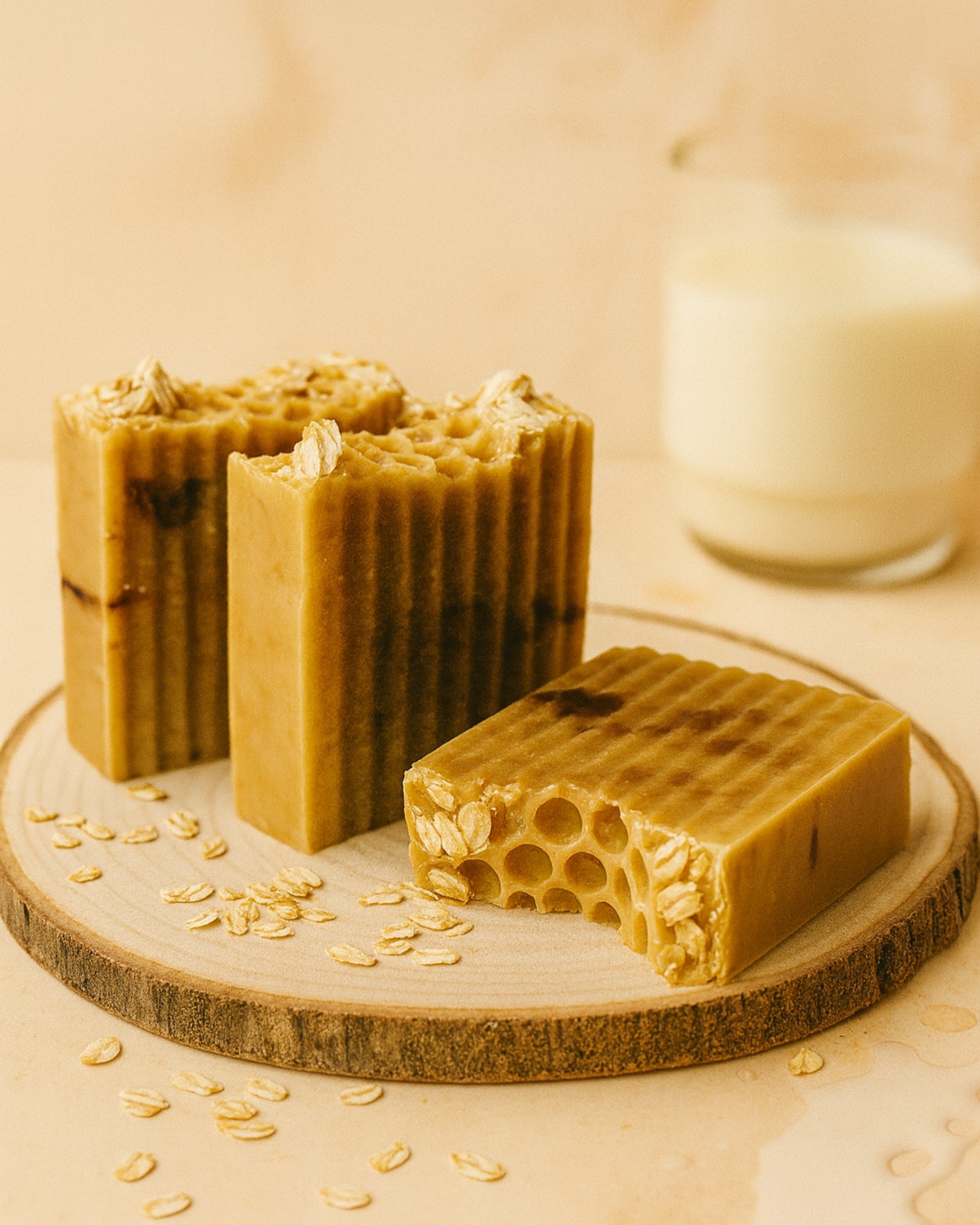Three bars of honeycomb-patterned soap on a wooden board with oats and a glass of oat milk in the background.