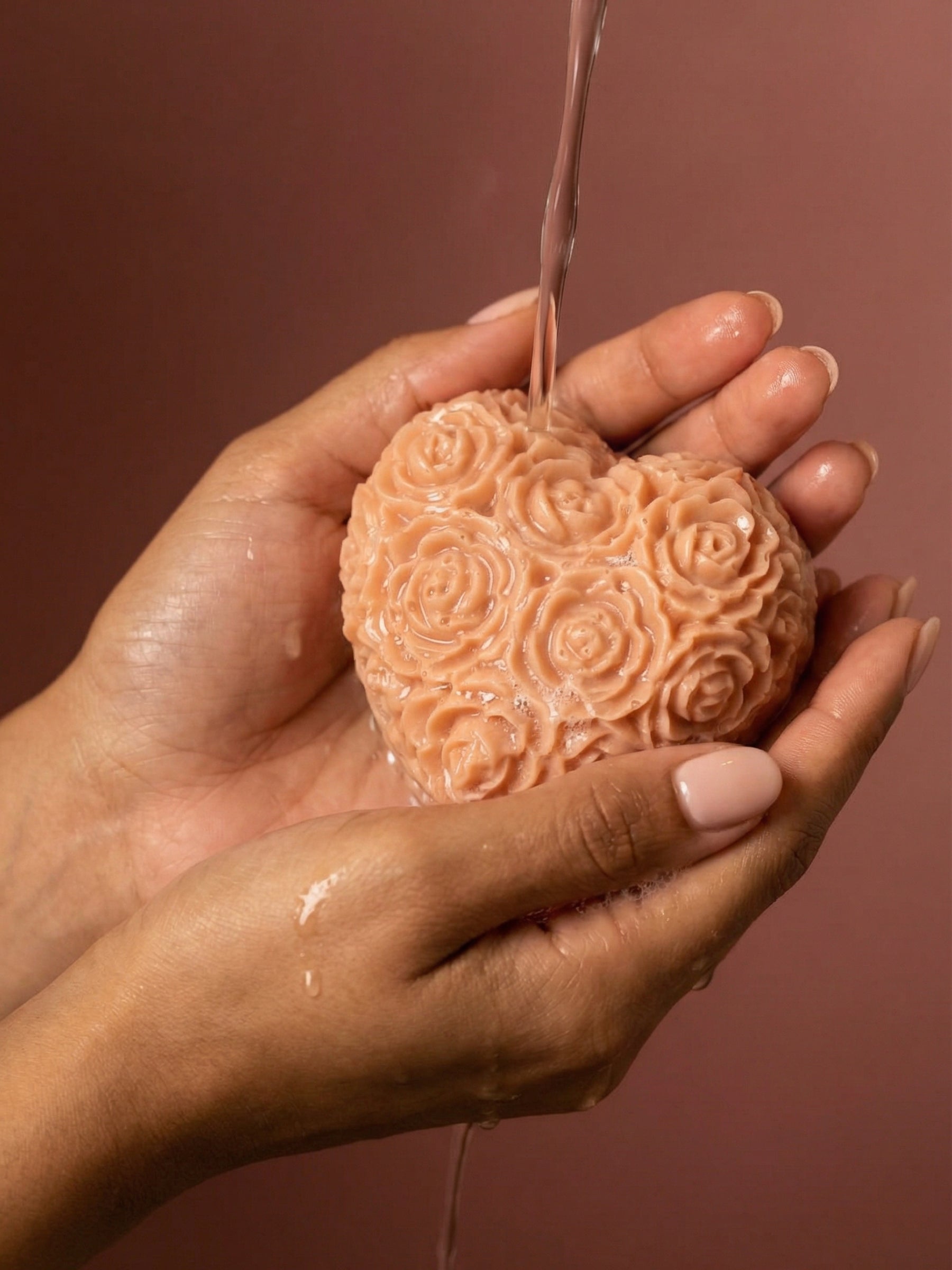 Heart-shaped The Fleur Grand Bloom rose-patterned rose geranium vanilla soap being held with water dripping from it against a brown background by DEOS CO