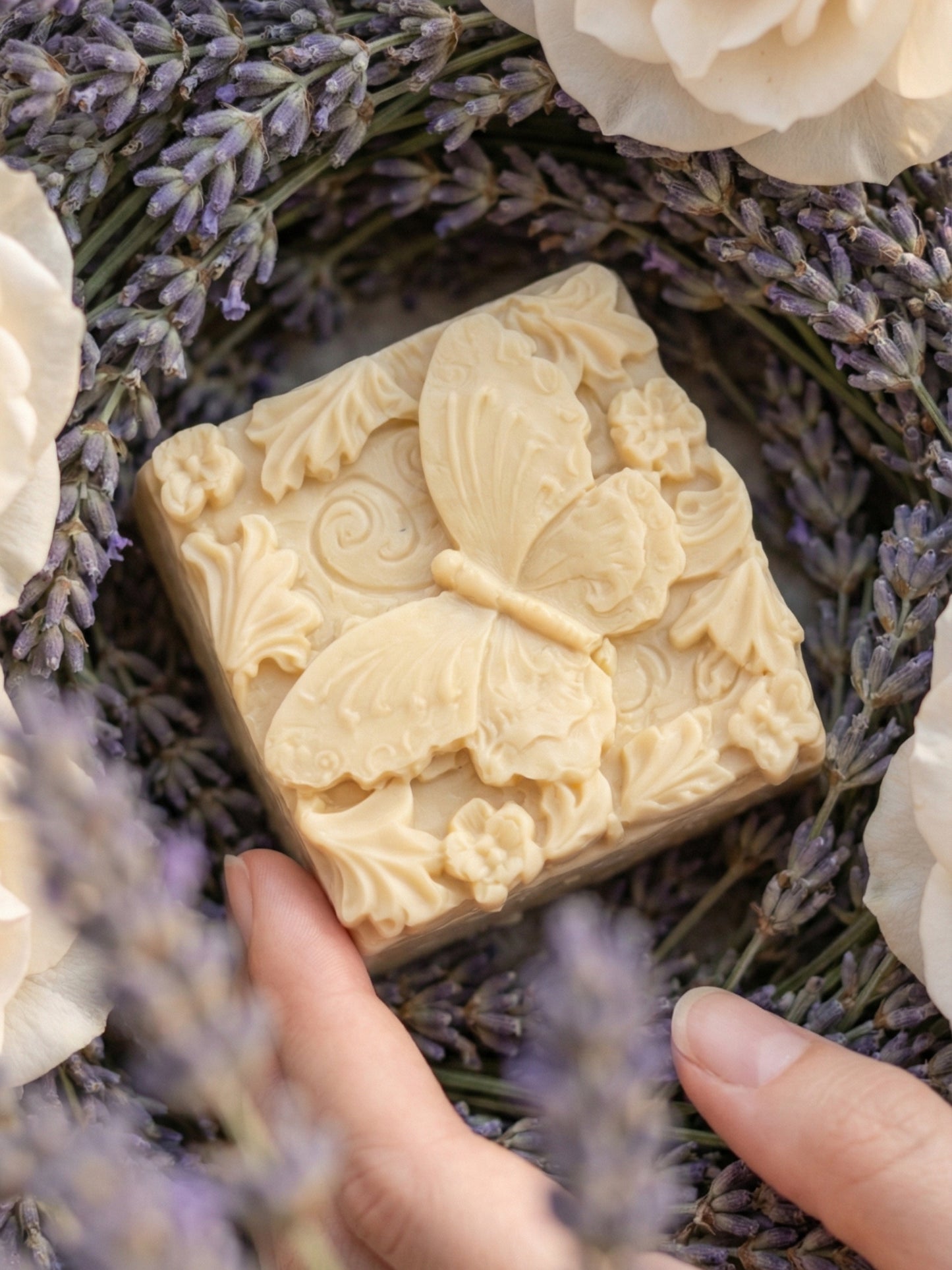 Butterfly-patterned olive oil soap bar held between lavender and white flowers.
The Metamorphé butterfly relief soap by DEOS Co., Nardus Bliss Collection—handcrafted with organic Greek olive oil and pure lavender essential oil, styled with dried lavender on textured stone.