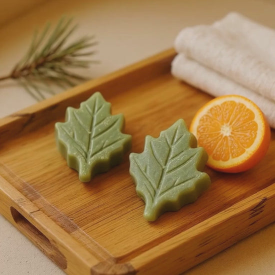Two leaf-shaped green soap bars on a wooden tray with an orange and a towel in the background.