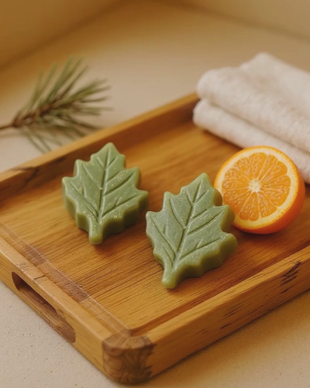 Two leaf-shaped green soap bars on a wooden tray with an orange and a towel in the background.
