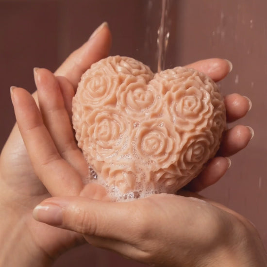 Heart-shaped The Fleur Grand Bloom rose-patterned rose geranium vanilla soap being held with water dripping from it against a brown background by DEOS CO