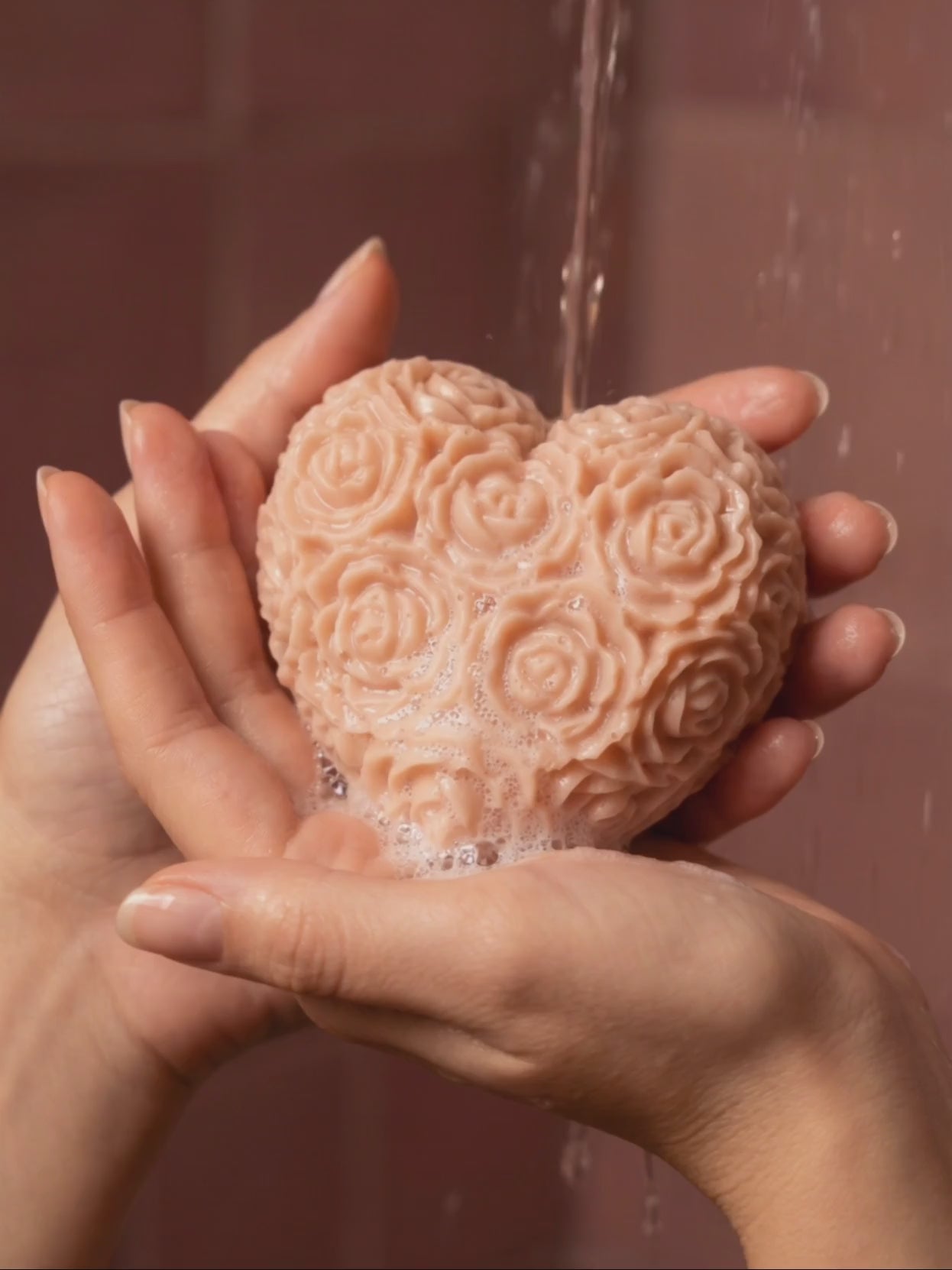 Heart-shaped The Fleur Grand Bloom rose-patterned rose geranium vanilla soap being held with water dripping from it against a brown background by DEOS CO